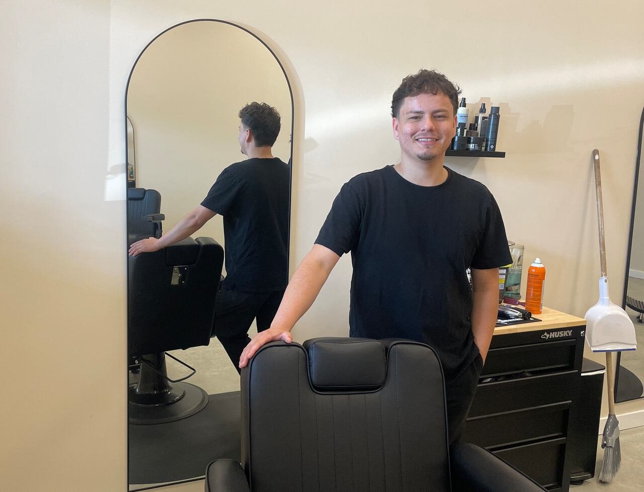 default-3175 A man in black attire stands next to a black barber chair with his hand on the back. Behind him, there is a long mirror and a black cart with hair tools and bottles.